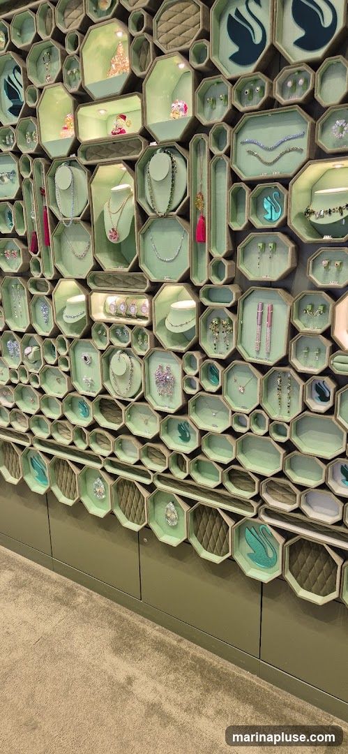 A wall display filled with various jewelry items, including necklaces, earrings, bracelets, and watches, arranged in hexagonal and octagonal compartments. Some compartments feature decorative elements like swan motifs and small figurines. The display is illuminated, highlighting the sparkling nature of the jewelry.