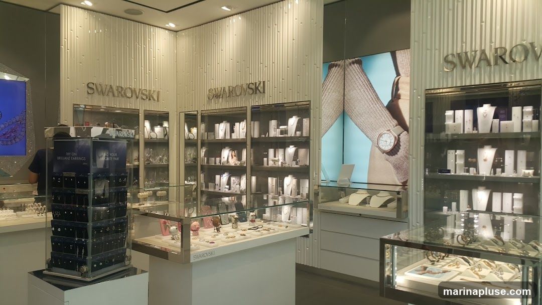 The interior of a Swarovski jewelry store, featuring display cases filled with necklaces, earrings, bracelets, and watches. The store has a clean, modern aesthetic with white walls and illuminated display shelves. A large advertisement featuring a woman wearing a watch is visible on the right side of the store.