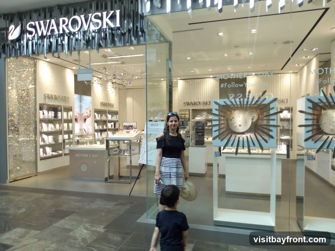 A woman and a child stand in front of a Swarovski store. The store has a large illuminated sign that reads "SWAROVSKI" above the entrance. The interior of the store is visible through the glass windows, displaying various jewelry items in display cases. There are also promotional displays for "Mother's Day" and a hashtag "#FollowYour".