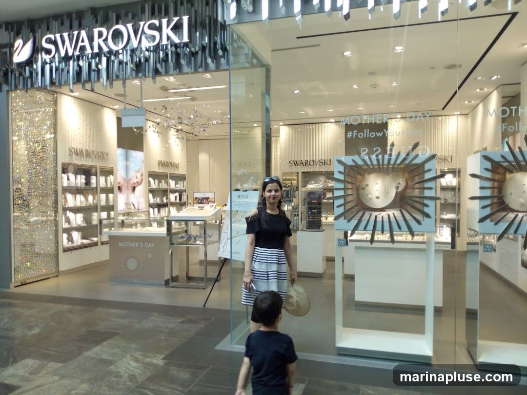 A woman and a child stand in front of a Swarovski store. The store has a large illuminated sign that reads "SWAROVSKI" above the entrance. The interior of the store is visible through the glass windows, displaying various jewelry items in display cases. There are also promotional displays for "Mother's Day" and a hashtag "#FollowYour".
