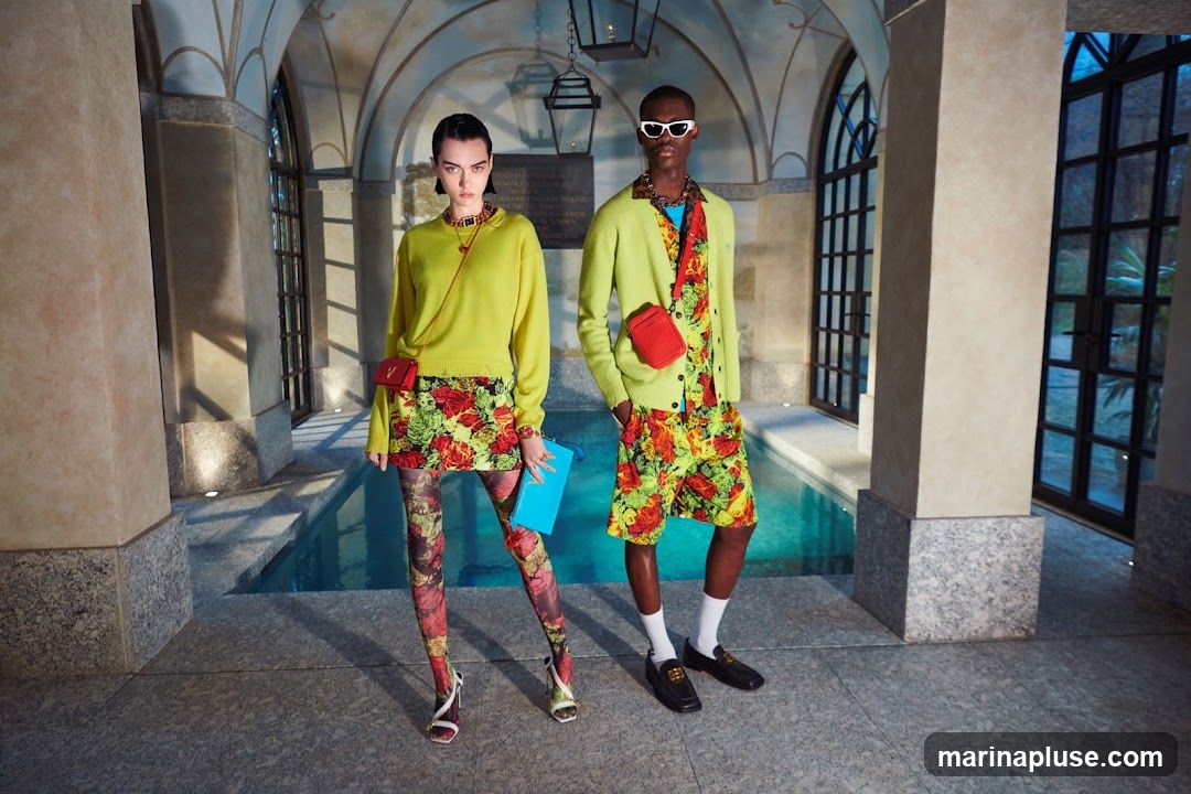 Two models, a woman and a man, stand in an indoor courtyard with a pool. The woman wears a lime green sweater, a floral print skirt, and patterned tights. She carries a blue clutch and a small red crossbody bag. The man wears a lime green cardigan over a floral print shirt and shorts. He also has a red crossbody bag. Both models wear sunglasses. The background features arched windows and stone architecture.