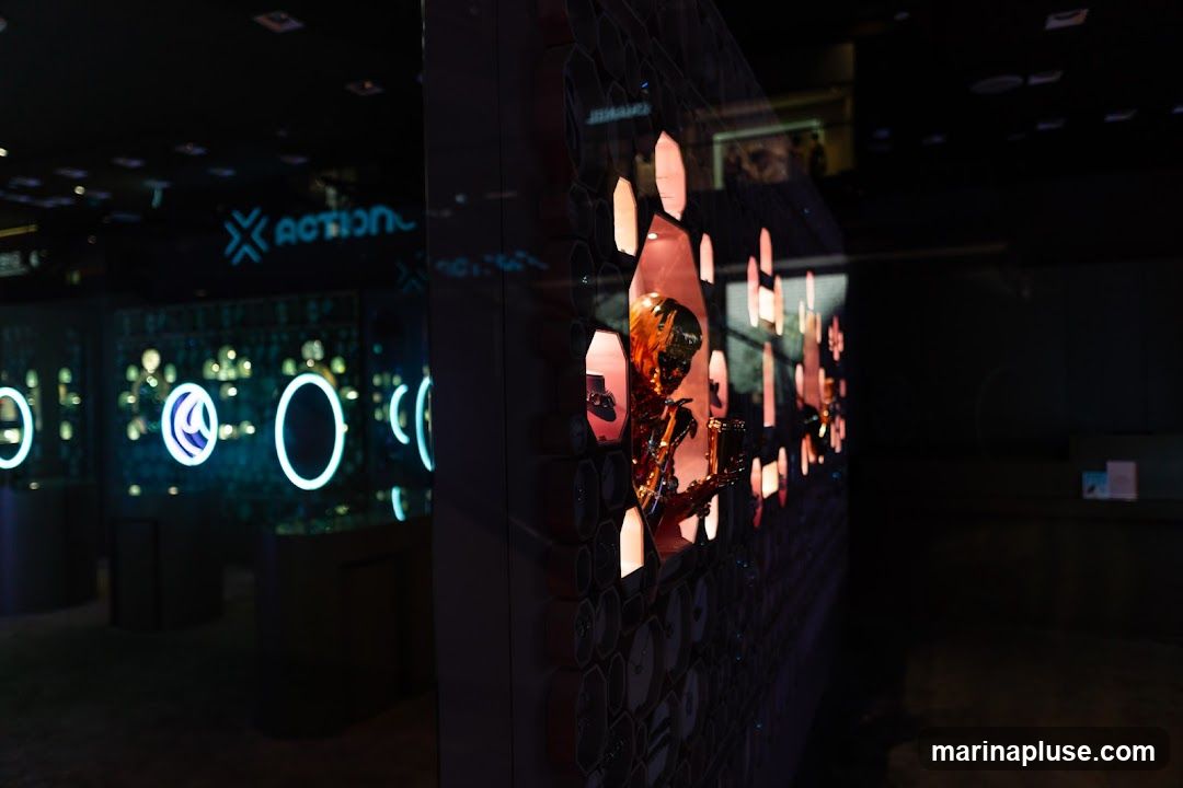 A store window display featuring a golden robot figure playing a saxophone, illuminated by pink and orange lights. In the background, a sign with the word "ACTION" in blue neon letters is visible, along with displays of watches or jewelry behind glass cases, accented by blue neon rings.