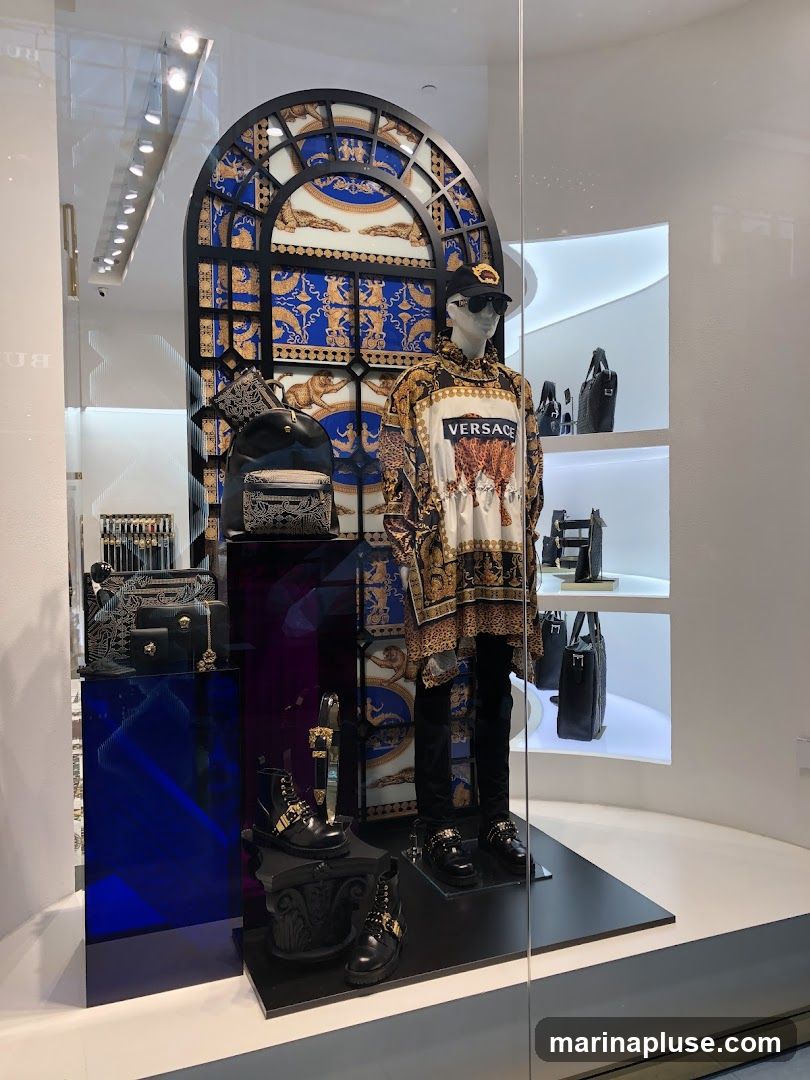 A store window display featuring a mannequin dressed in a Versace outfit, including a patterned shirt with the brand name, black pants, a cap, and sunglasses. The display also showcases various Versace accessories such as backpacks, handbags, wallets, and boots. The background is decorated with a large, ornate archway featuring blue and gold patterns.