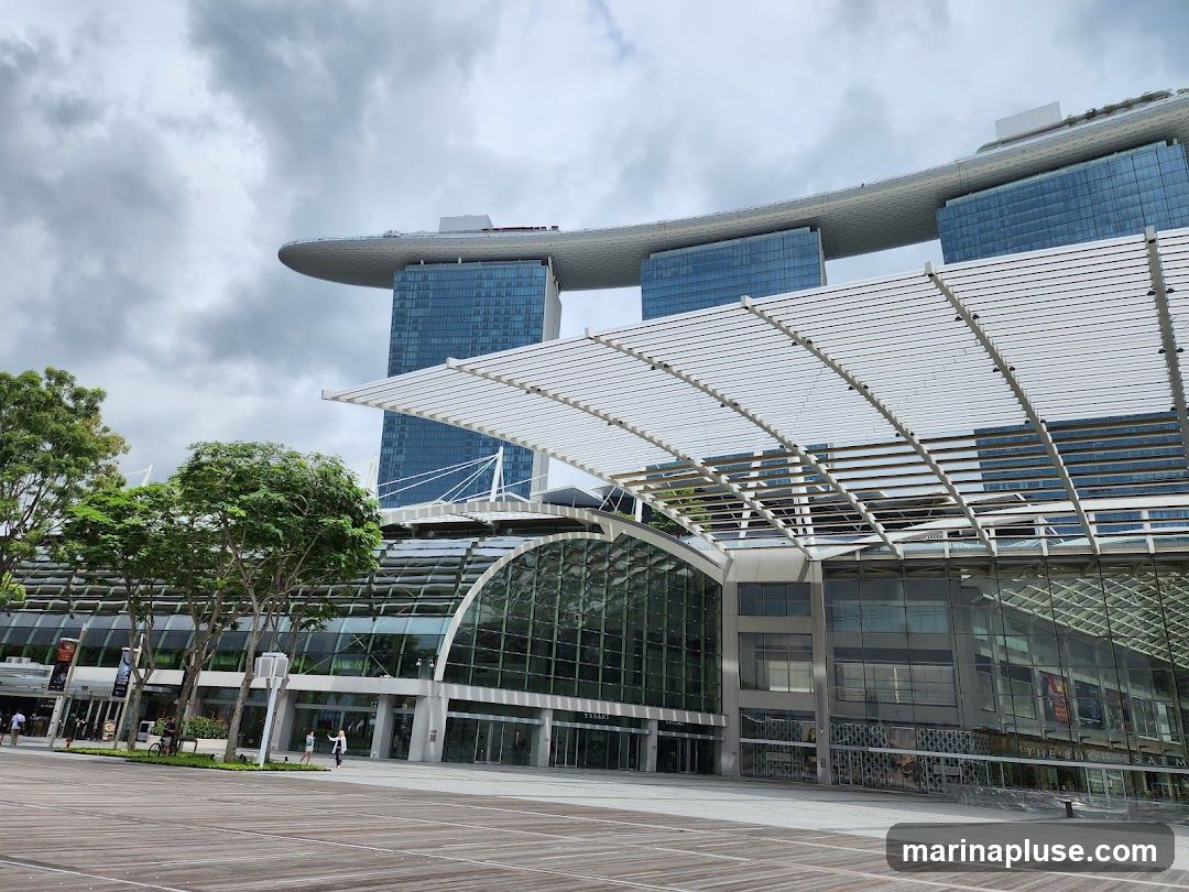 A wide shot of the Marina Bay Sands complex in Singapore under a cloudy sky. The iconic three towers with the ship-like structure on top are visible in the background. In the foreground, the modern architecture of the convention center or shopping mall is prominent, featuring a large glass facade and a white slatted roof structure. Trees and a wooden walkway are also visible.
