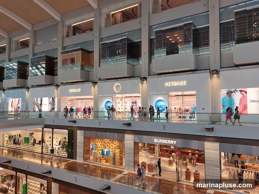 A wide shot of the interior of a multi-level luxury shopping mall. Several high-end stores are visible, including Versace and Burberry, with their prominent signage. People are seen browsing and walking along the elevated walkways. The architecture features modern design with glass facades and clean lines.