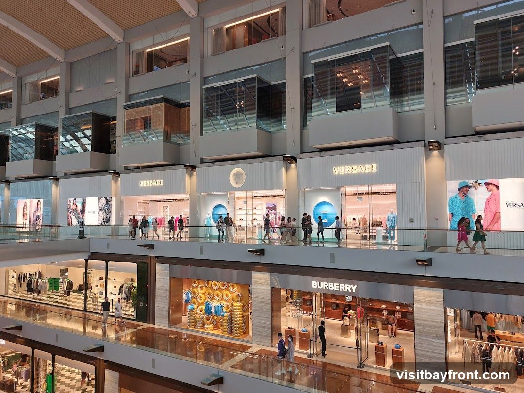 A wide shot of the interior of a multi-level luxury shopping mall. Several high-end stores are visible, including Versace and Burberry, with their prominent signage. People are seen browsing and walking along the elevated walkways. The architecture features modern design with glass facades and clean lines.