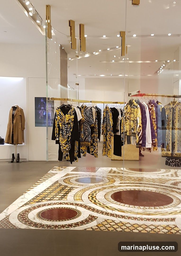 The image shows the interior of a high-end fashion boutique. Racks of clothing, featuring bold patterns and vibrant colors, are displayed. A mannequin on the left is dressed in a tan trench coat and black boots. The floor is adorned with an intricate mosaic pattern. The overall aesthetic suggests a luxurious and stylish retail environment.