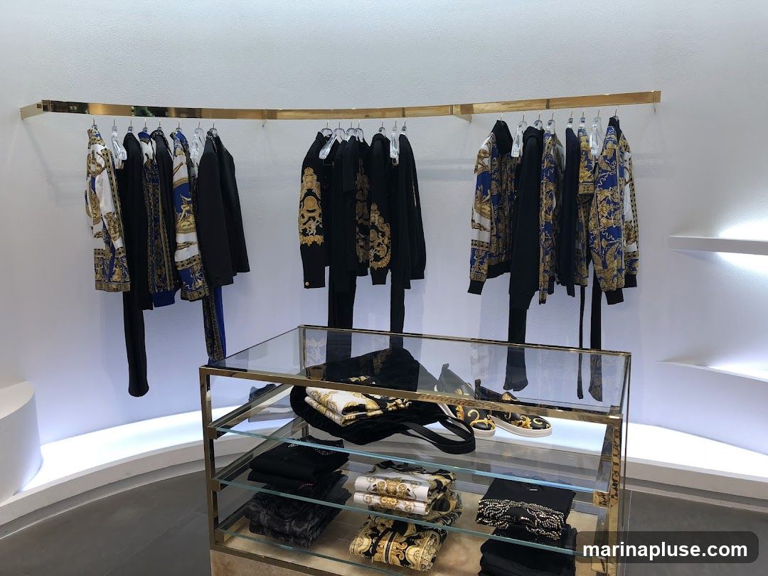 A display of luxury clothing, featuring jackets and pants with a distinctive blue and gold baroque pattern, hung on a curved gold rack. Below, a glass display case showcases folded garments, a handbag, and shoes, also in the same ornate style. The store interior is minimalist with white walls and subtle lighting.