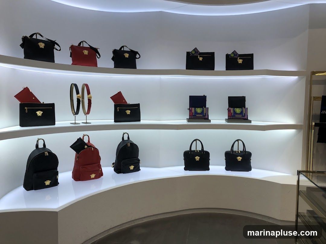 A well-lit display shelf showcases a collection of luxury handbags and accessories. The items are arranged on white, curved shelves with integrated lighting. On the top shelf, there are three handbags, two black and one red, each adorned with a gold Medusa head emblem. The middle shelf features black clutches with gold hardware, a red clutch, and two decorative belts with the same emblem. The bottom shelf displays three backpacks, two black and one red, and two black handbags with top handles, all featuring the signature gold Medusa head.