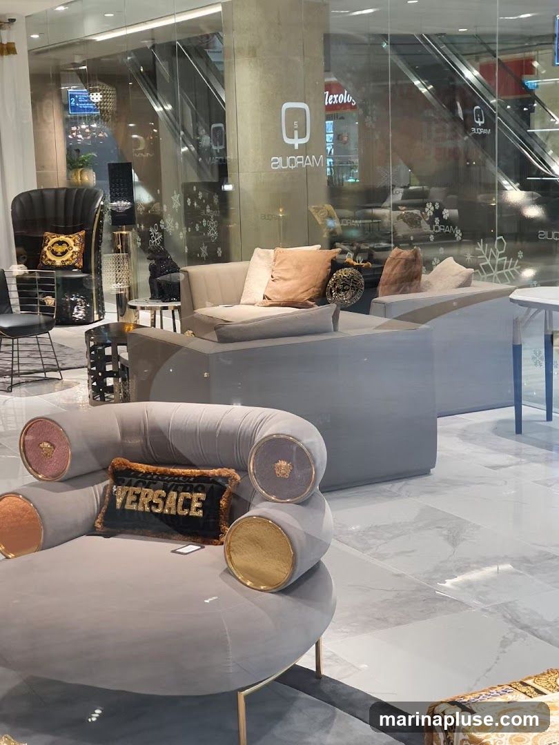 A display of high-end furniture, including a grey sectional sofa with accent pillows and a unique grey armchair with gold and circular accents. A black pillow with the word "VERSACE" in gold lettering is placed on the armchair. The display appears to be inside a store, with reflections of escalators and other shops visible in the glass storefront.