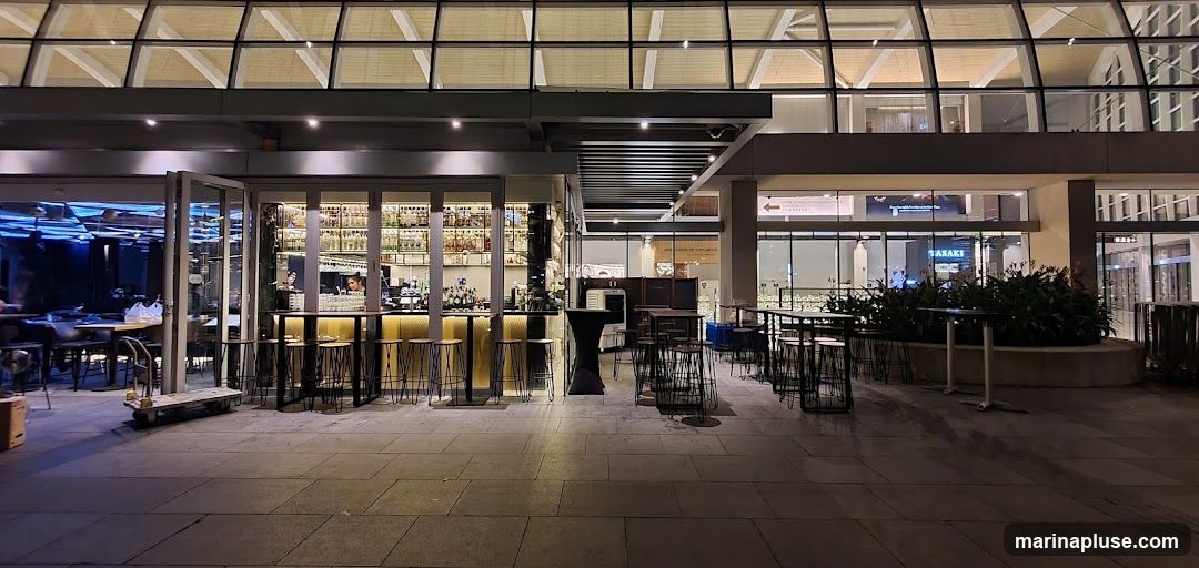 An outdoor seating area in front of a bar with a large glass facade. The bar is well-lit with bottles displayed on shelves behind the counter. Several high-top tables with stools are arranged outside, some with umbrellas. The overall ambiance is modern and inviting, suitable for evening dining or drinks.