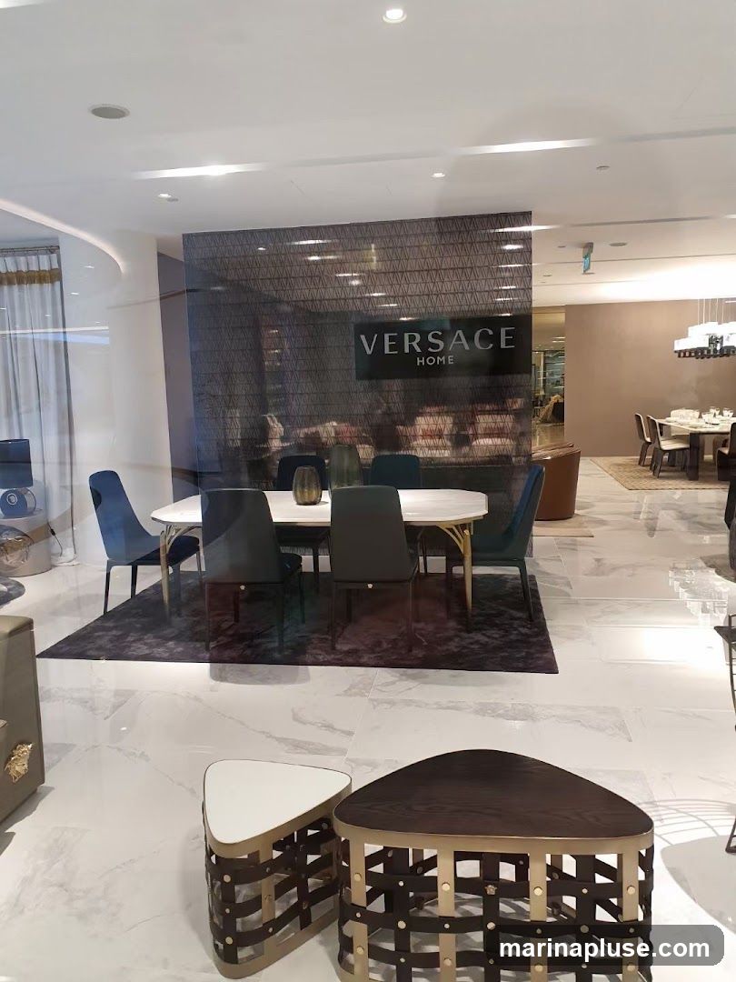 A well-lit showroom displays a dining table with chairs, coffee tables, and other furniture. The 'VERSACE HOME' logo is visible on a patterned wall in the background.