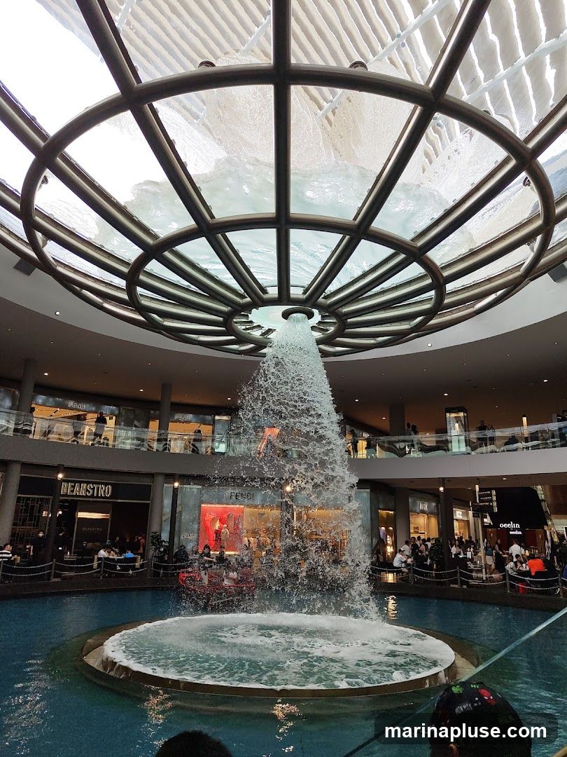 An indoor fountain cascades water from a circular structure in the ceiling into a pool below. Surrounding the pool are various shops and restaurants on multiple levels, with people visible inside and outside the establishments. Brands like Fendi and Beanstro are identifiable.