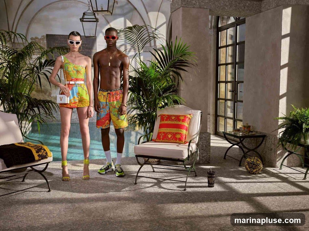 A male and female model stand by a pool, showcasing vibrant Versace swimwear. The female model wears a one-shoulder top and shorts set with a white belt and a small white handbag. The male model wears patterned swim shorts. They are surrounded by lush green plants and elegant outdoor furniture, including chairs with decorative pillows and a small table with a tea set. The scene is set in a luxurious, sun-drenched interior with arched windows and stone flooring.