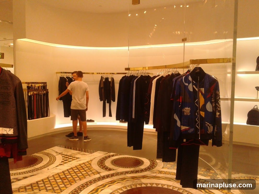 The image shows the interior of a high-end clothing store. Racks of men's clothing, including jackets, shirts, and pants, are displayed. A display of belts is visible on the left. Two men are browsing the merchandise. The store features a modern design with curved walls, ambient lighting, and a decorative marble floor.