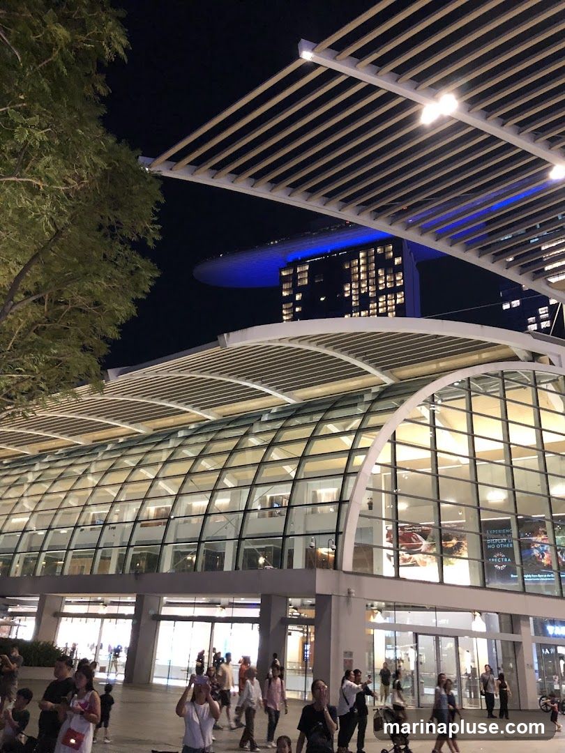 A modern building with a curved glass facade is illuminated at night. People are walking in front of the building. In the background, a tall building with a distinctive boat-like structure on top is visible, lit with blue lights.