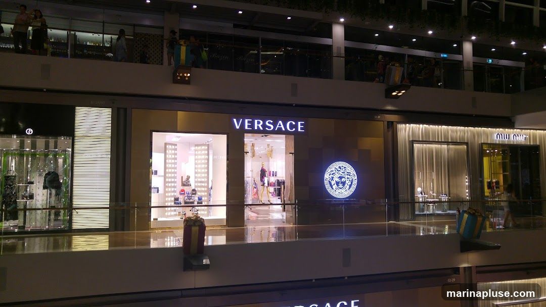 The exterior of a Versace store in a mall. The store has a large illuminated Versace logo and the word "VERSACE" displayed prominently above the entrance. Inside, mannequins are visible displaying clothing. To the left is another store with a logo that appears to be for Giorgio Armani, and to the right is a Miu Miu store.