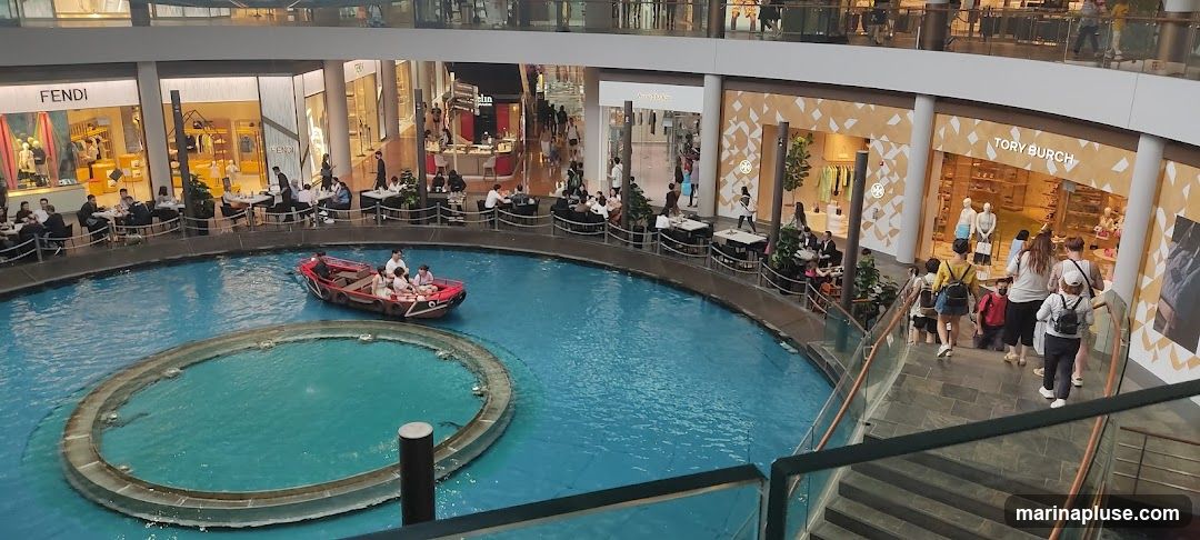 An aerial view of a bustling indoor shopping mall featuring a canal with a boat carrying passengers. Surrounding the canal are various shops, including Fendi and Tory Burch, with people dining at tables set up along the walkway. The architecture is modern, with multiple levels visible.