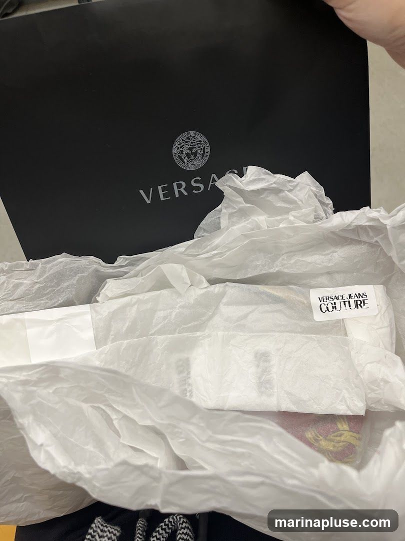 A black Versace shopping bag with the brand's logo and name is visible at the top of the image. Below the bag, white tissue paper is used to package items, with a label that reads "VERSACE JEANS COUTURE" visible on one of the items.