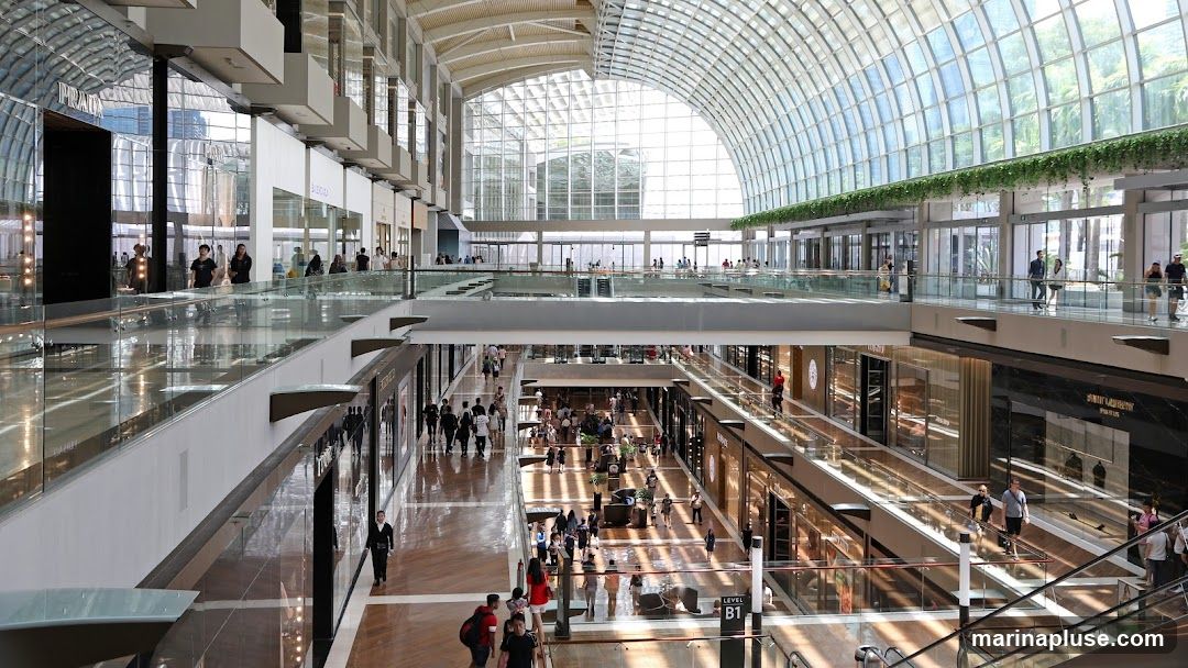 A wide shot of the interior of a modern, multi-level shopping mall. Sunlight streams in through a large, arched glass ceiling, illuminating the polished wooden floors and glass railings of the walkways. People are seen browsing in various stores and walking along the different levels. Storefronts for luxury brands like Prada and Saint Laurent are visible.