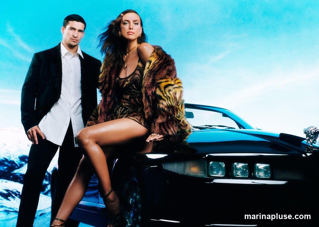 A fashion editorial image featuring a male model in a black suit and white shirt standing next to a female model. The female model is seated on the hood of a black car, wearing a tiger-print faux fur coat over a patterned dress and high heels. The background is a bright blue sky with wispy clouds and a hint of a mountainous landscape.