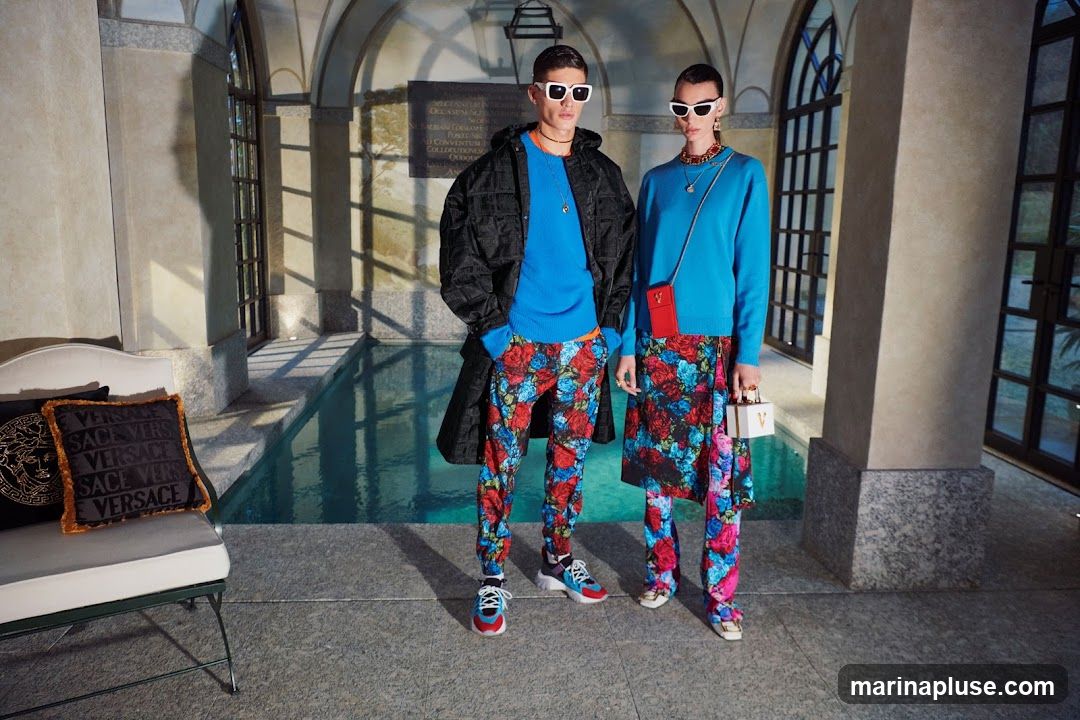 Two models, a man and a woman, stand near a pool. The man wears a black jacket over a blue sweater and floral pants. The woman wears a blue sweater, a floral skirt, and floral pants. Both wear white sunglasses and jewelry. A white couch with Versace-branded pillows is visible on the left.