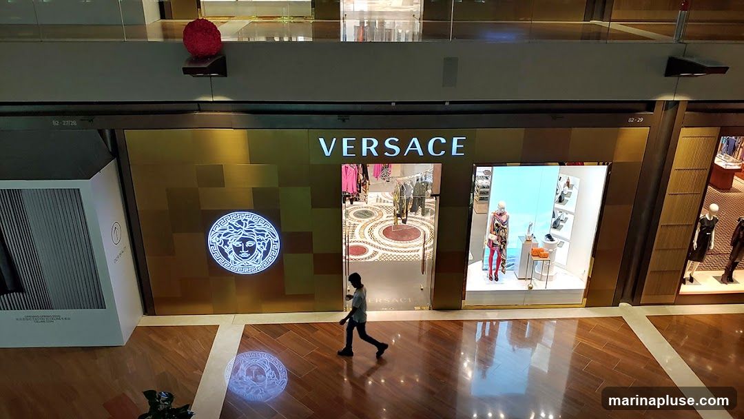 The exterior of a Versace store is shown, featuring the brand's name in large white letters above the entrance. To the left of the entrance is a large, illuminated circular logo of the Medusa head. The store has a golden facade with a geometric pattern. Through the glass entrance, racks of clothing and mannequins displaying merchandise are visible. To the right of the entrance, a display window showcases mannequins dressed in fashionable outfits. The floor is made of polished wood, reflecting the store's interior and signage. A person is walking in front of the store.