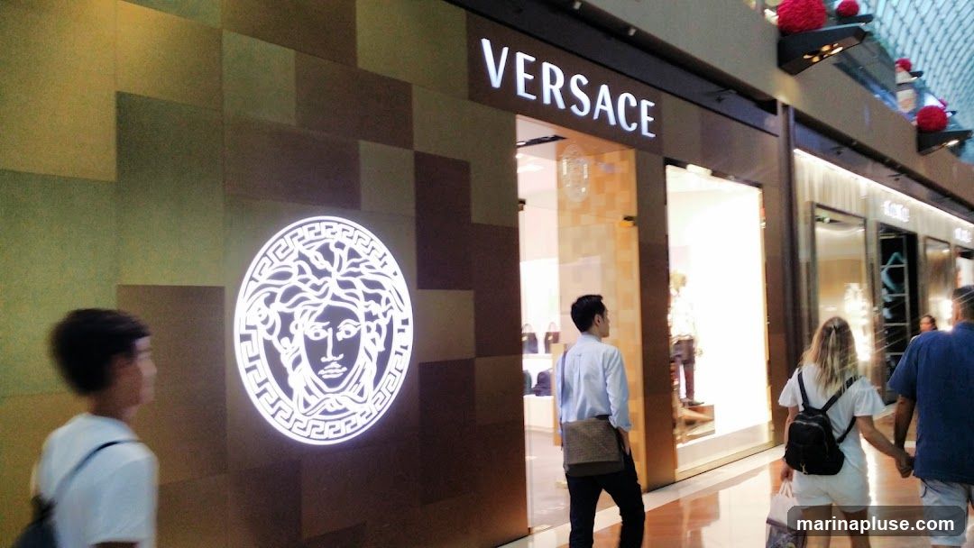 The exterior of a Versace store with its iconic Medusa logo illuminated on the left and the "VERSACE" name displayed above the entrance. People are walking past the store.