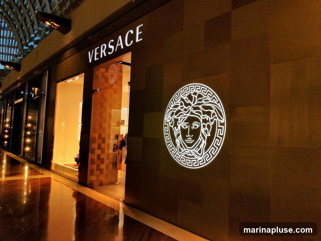 The exterior of a Versace store at night. The store has a large glass entrance and a prominent illuminated Versace logo on the dark facade. The word "VERSACE" is displayed in illuminated letters above the entrance.