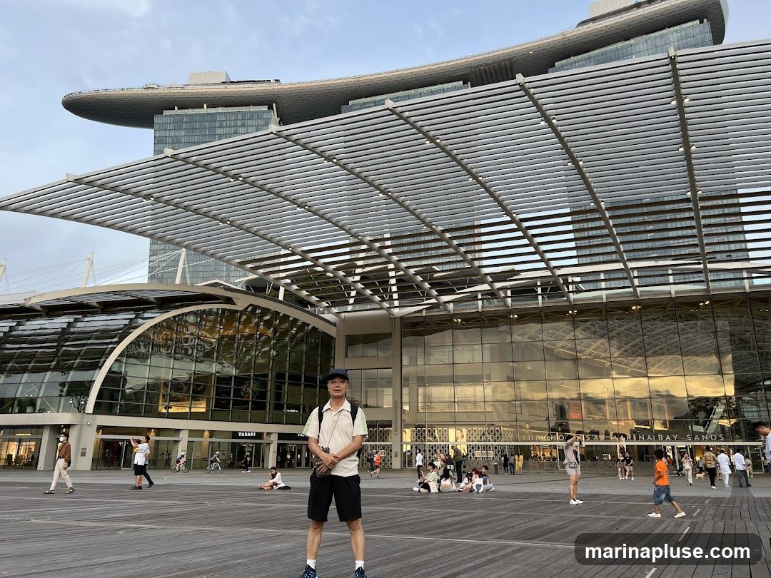 A man stands in the foreground on a wooden boardwalk, facing the camera. Behind him is the exterior of the Marina Bay Sands complex, with its distinctive architecture. Several other people are scattered around the plaza, some sitting and some walking. The sky is overcast.