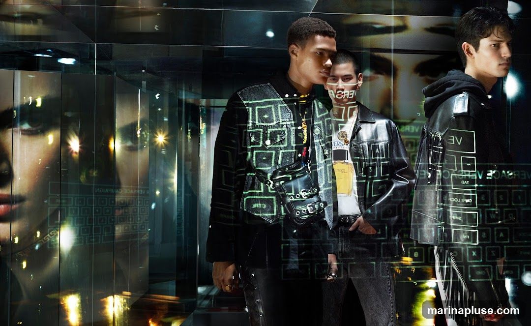 Three male models are showcased in a visually striking setting with reflective surfaces and digital overlays. The models are dressed in contemporary fashion, featuring leather jackets, denim, and accessories like bags and necklaces. Digital graphics, including what appears to be a keyboard interface and text, are projected or reflected onto the scene, creating a futuristic and high-fashion aesthetic. A bottle of Versace Eros perfume is visible on one of the models' shirts.