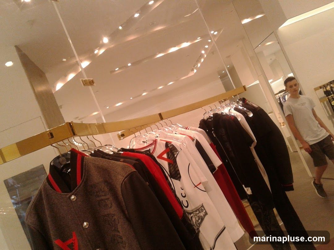 A young man stands in a brightly lit clothing store, looking into a mirror. Racks of men's clothing, including jackets and t-shirts, are displayed in the foreground. The store features a modern design with mirrored walls and ceiling lights.