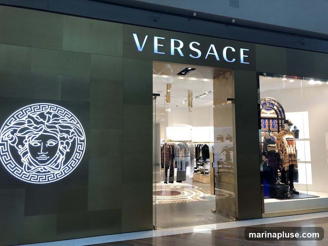 The exterior of a Versace store. The store has a large, illuminated Medusa logo on the left side of the facade and the word "VERSACE" in large, illuminated letters above the entrance. The entrance is a glass doorway, and the store windows display mannequins dressed in Versace clothing and accessories. The interior of the store is visible through the glass, showing racks of clothing and display shelves.
