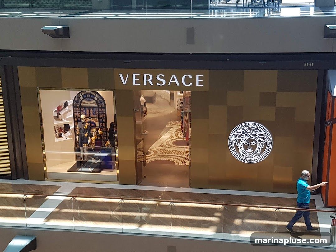 A wide shot of the Versace store entrance. The store has a large gold facade with the "VERSACE" logo in white lettering above the entrance. To the left of the entrance is a display window showcasing handbags and clothing on mannequins. To the right of the entrance is the iconic Versace Medusa head logo. A man in a blue shirt and dark pants is walking past the store.