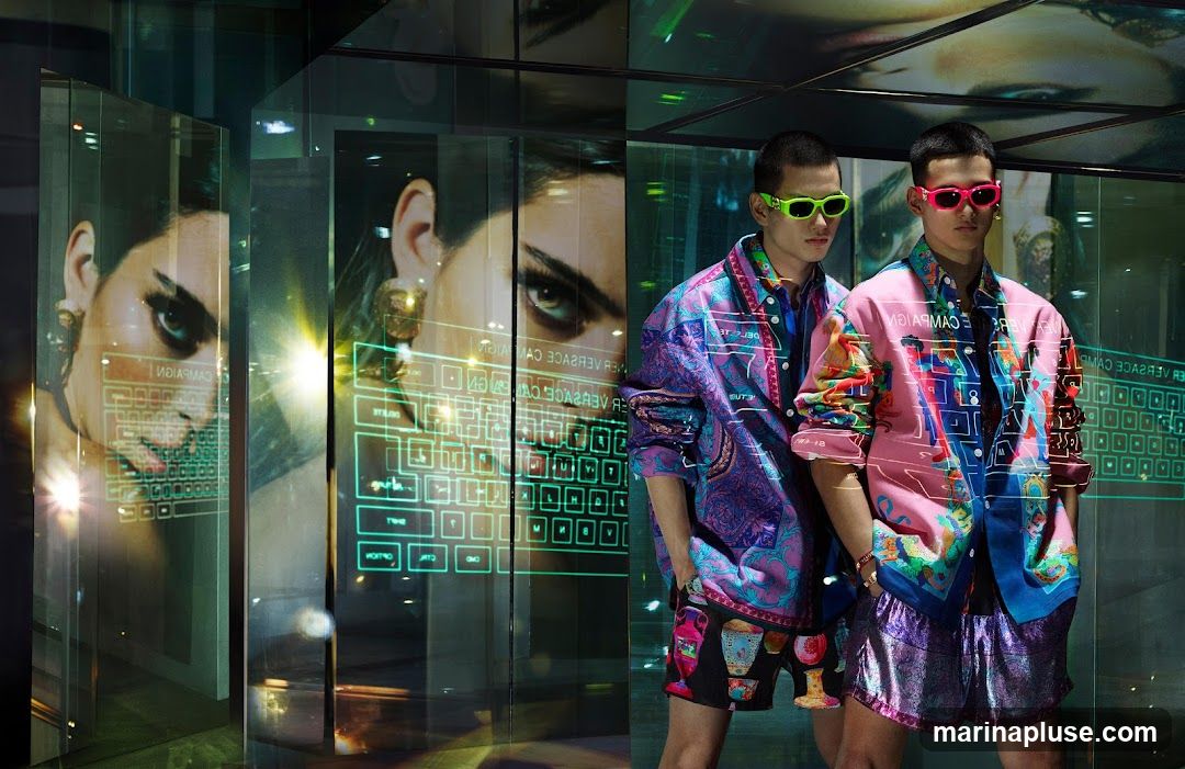 Two male models stand in front of a display featuring large images of a woman's face and a digital keyboard interface. The models are wearing colorful, patterned Versace shirts and shorts. The display appears to be a shop window or an interior promotional setup.