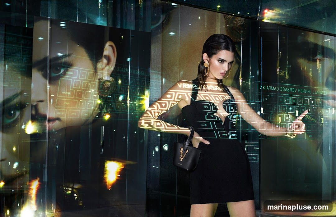 A model, identified as Kendall Jenner, poses in a black dress and holds a black handbag with a gold logo. She is surrounded by digital projections of a keyboard and a large image of her face. The background appears to be a reflective surface, possibly glass, showing city lights and reflections. The overall aesthetic is futuristic and high-fashion.