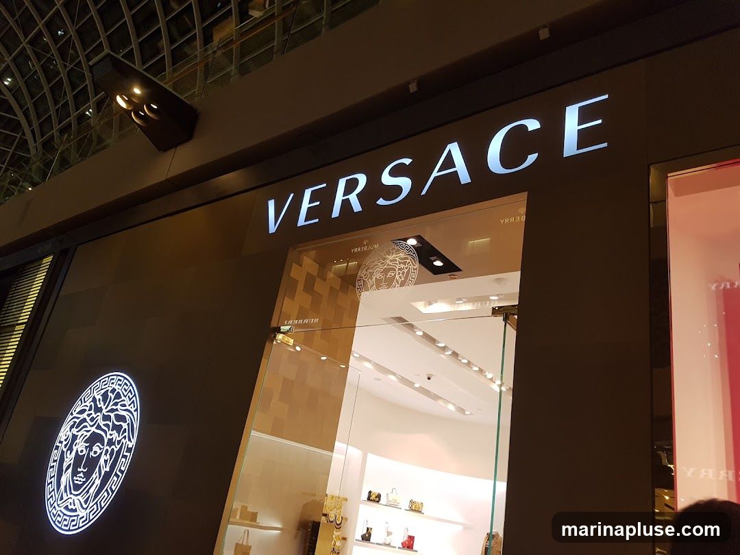 The exterior of a Versace store is shown, with the brand name "VERSACE" illuminated in large white letters above the entrance. To the left of the entrance, the iconic Versace Medusa head logo is displayed within a circular emblem, also illuminated. The entrance features glass doors and windows, offering a glimpse into the store's interior where handbags and other merchandise are displayed on shelves. The overall impression is one of luxury and high fashion.
