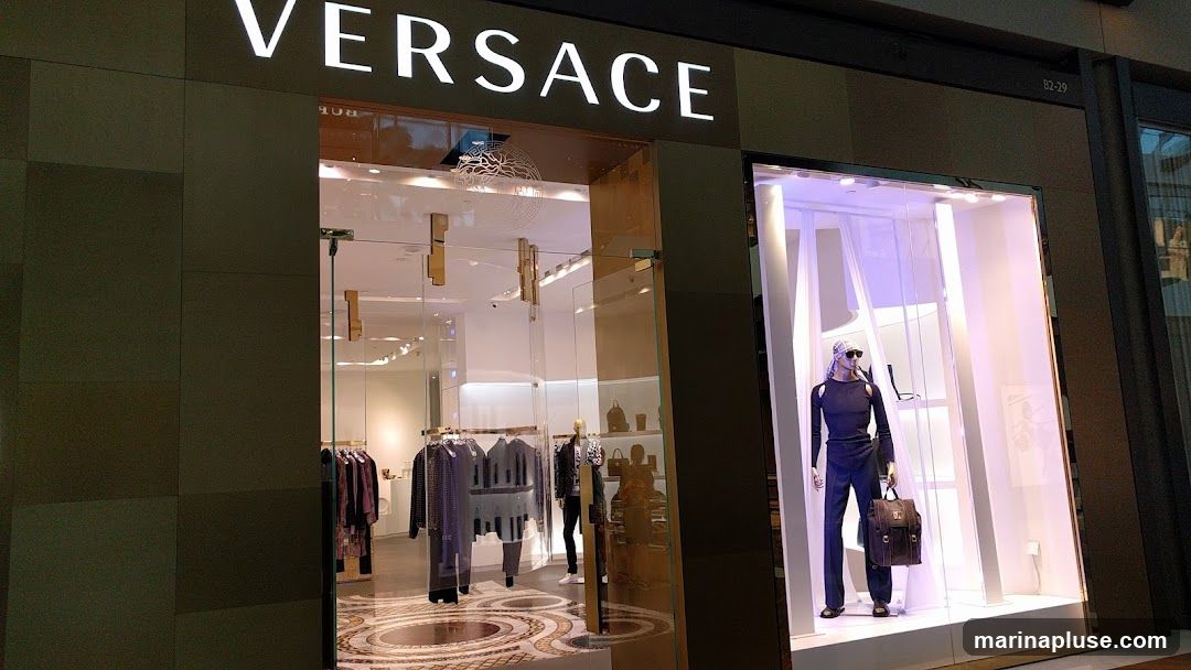 The exterior of a Versace store with a large illuminated sign above the entrance. The entrance is a glass door with gold handles, leading into a well-lit interior displaying clothing on racks and mannequins. To the right, a display window features a mannequin dressed in a dark outfit, accessorized with sunglasses and a brown leather bag. The store's facade is dark, contrasting with the bright interior and display window.