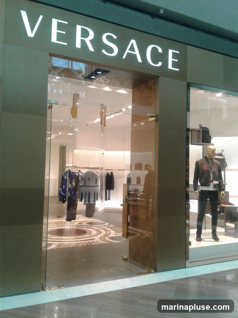 The exterior of a Versace store with the brand name prominently displayed in large white letters above the entrance. The store features a glass facade, allowing a view of the interior displays of clothing and accessories. A mannequin dressed in a jacket and pants is visible in the right-hand display window.