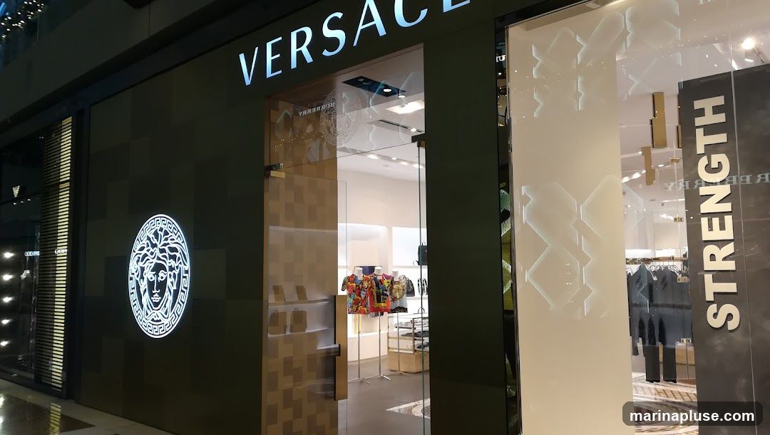 The exterior of a Versace store. The store has a dark green facade with the Versace logo prominently displayed. The entrance is a glass door, and through the glass, mannequins displaying colorful clothing and shelves with handbags can be seen. To the right of the entrance, there is a vertical sign that reads "STRENGTH" and the Burberry logo is visible on the glass.