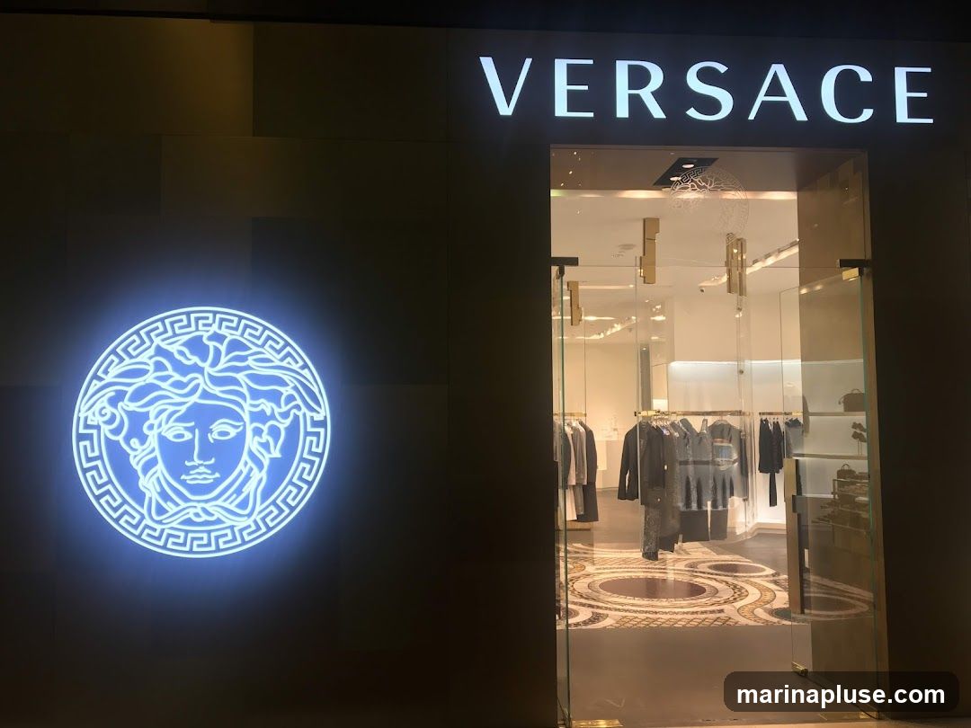 The exterior of a Versace store is shown at night. A large, illuminated blue Medusa logo is visible on the dark facade to the left. To the right, the store entrance features glass doors and windows, revealing a well-lit interior with clothing racks displaying garments. The word "VERSACE" is illuminated in white letters above the entrance.