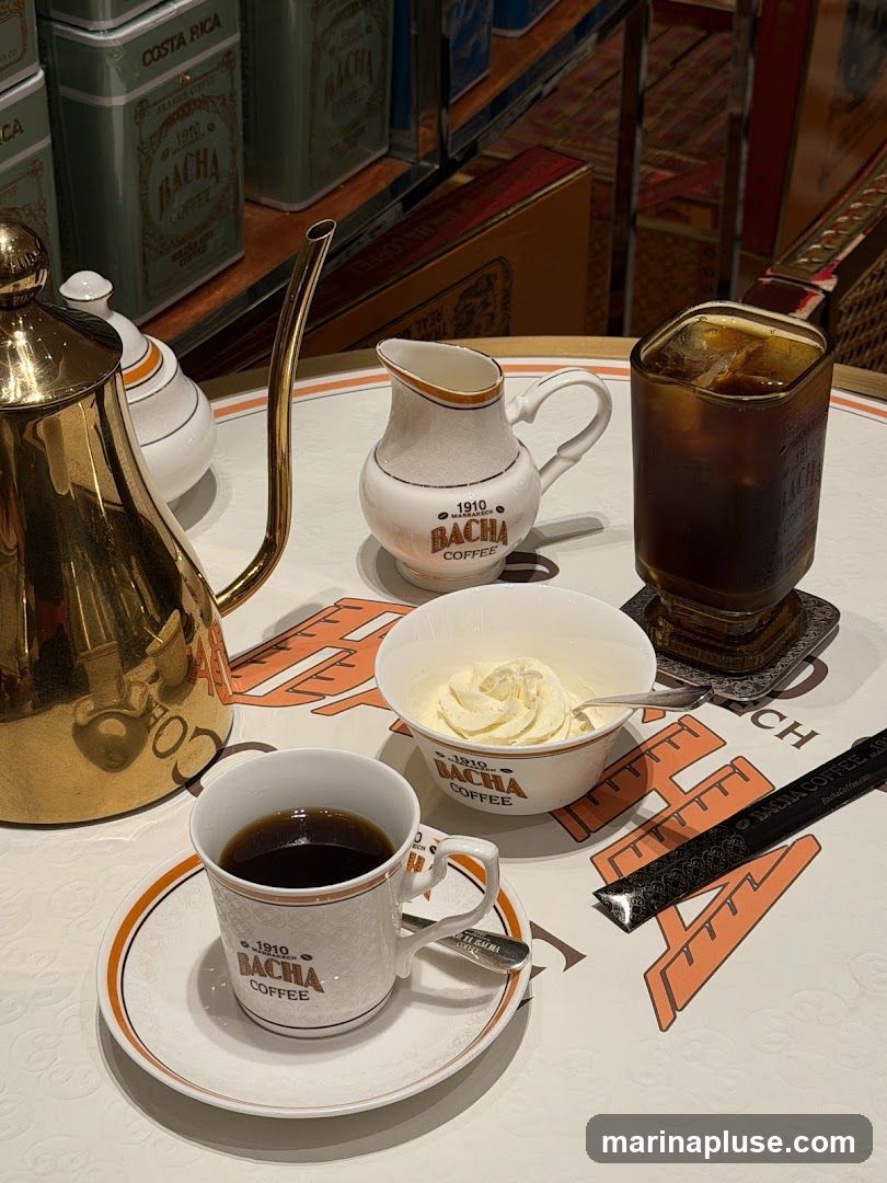 A table setting with a gold coffee pot, a cup of black coffee, a small pitcher of cream, a bowl of whipped cream, and a glass of iced coffee. The tableware is branded with "1910 BACHA COFFEE". In the background, there are shelves with branded coffee tins.