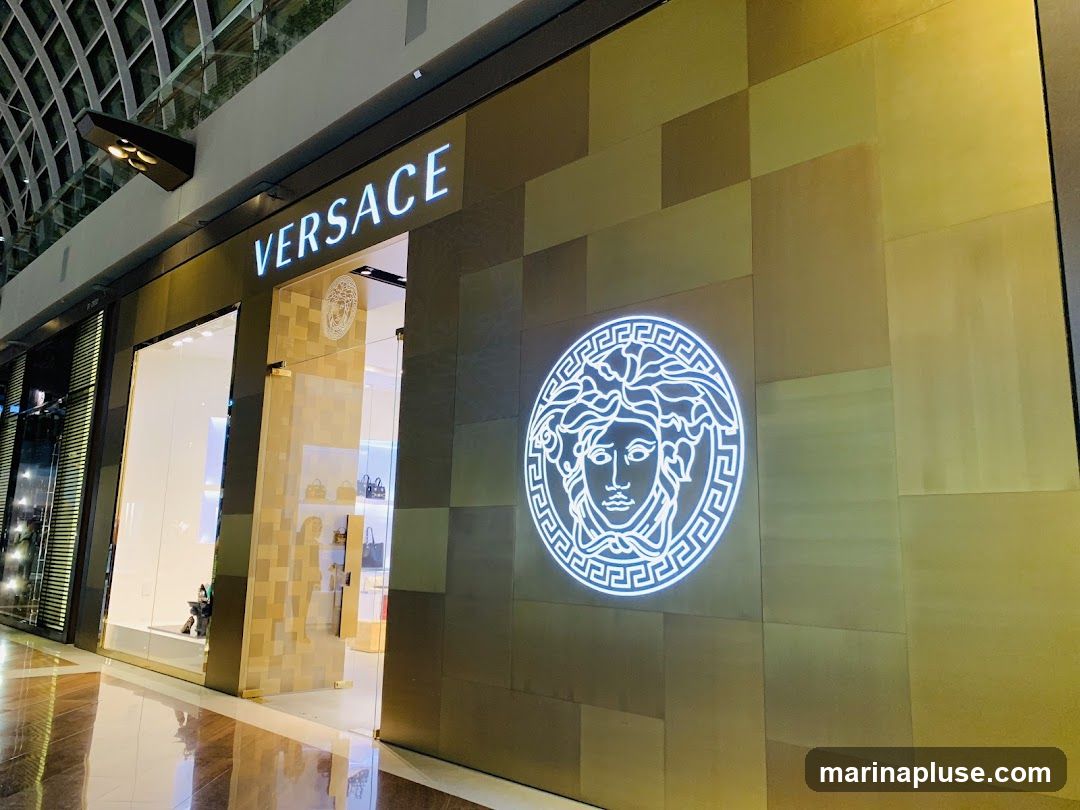 The exterior of a Versace store with the brand name illuminated above the entrance. A large, illuminated Medusa logo is displayed on the facade. The store features large glass windows and doors, with handbags visible inside. The facade is covered in gold-toned tiles.