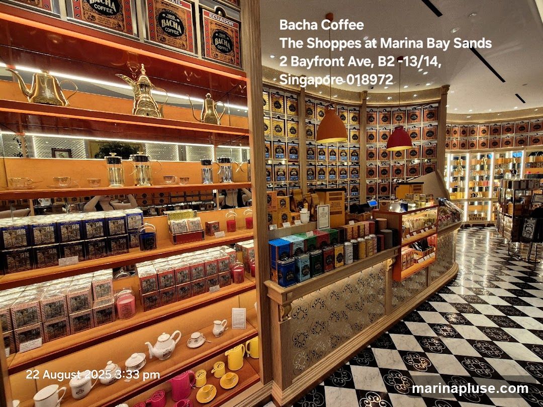The interior of a Bacha Coffee store, featuring shelves stocked with various tea and coffee products in decorative tins and boxes. Golden teapots and coffee makers are displayed on higher shelves. The store has a checkered black and white floor and warm lighting. The text overlay indicates the store is located at The Shoppes at Marina Bay Sands in Singapore.