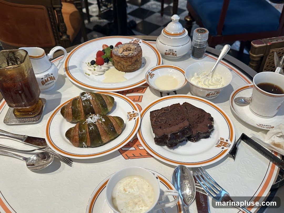 A table laden with various desserts and beverages, including two green croissants topped with shredded coconut, a slice of chocolate cake, a pastry with berries and cream, a glass of iced coffee, a cup of coffee, and a cup of what appears to be affogato. Branded tableware with "1910 BACHA COFFEE" is visible.
