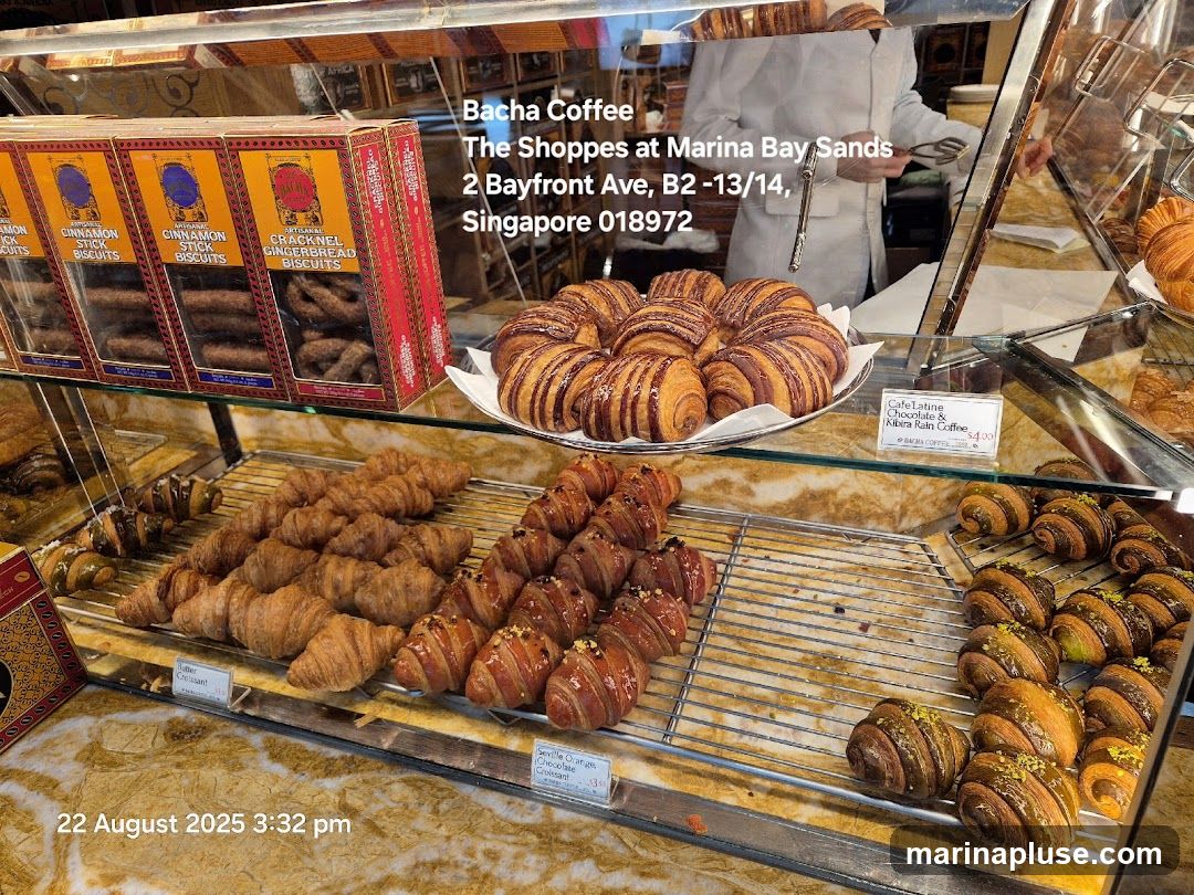 A display case filled with various pastries and biscuits from Bacha Coffee. In the foreground, there are rows of croissants, some plain, some chocolate-glazed, and some with pistachio toppings. Behind them, boxes of biscuits are stacked, labeled "Cinnamon Stick Biscuits" and "Cracknel Gingerbread Biscuits." A sign indicates "Cafe Latine Chocolate & Kibira Rain Coffee $4.00." The shop's name and address are visible on a sign in the background.