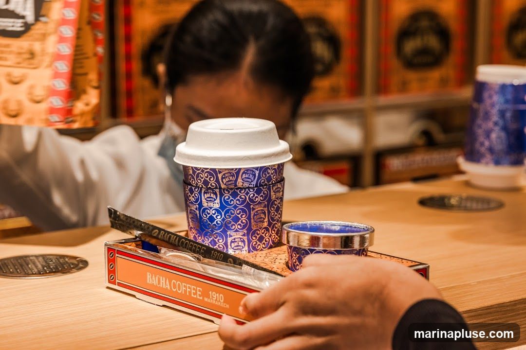 A person wearing a mask and a white shirt is preparing a coffee order. In the foreground, there is a branded coffee cup with a blue and gold pattern, a small blue cup, and a box labeled "BACHA COFFEE 1910 MARRAKECH" containing what appears to be sugar packets and a stirrer. The background shows shelves stocked with more branded coffee cups and packaging.