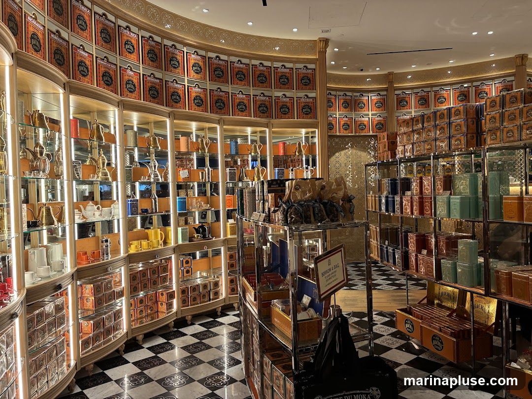 The interior of a luxurious tea shop, featuring shelves stocked with numerous decorative tins of tea. The walls are lined with rows of these tins, and display cases showcase teapots, cups, and other tea-related accessories. The floor has a distinctive black and white checkered pattern. A sign that reads "BORROW A BAG" is visible on a display stand.