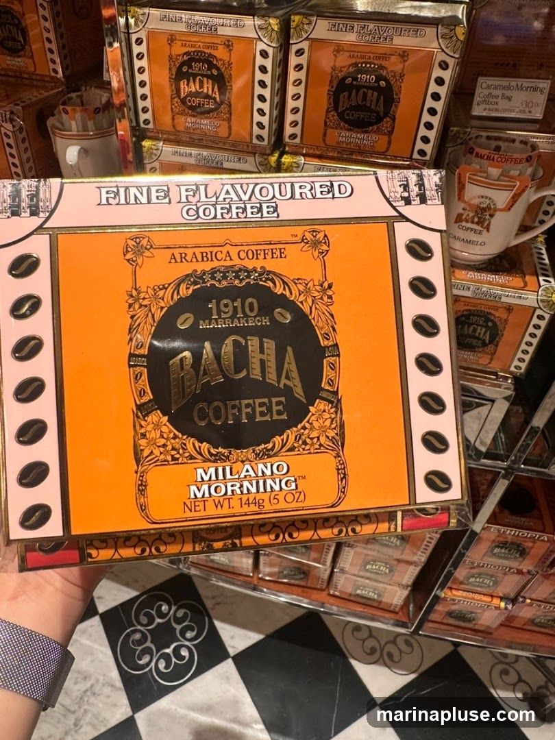 A close-up shot of Bacha Coffee products, featuring several boxes of fine flavored coffee. The prominent box in the foreground is orange with black and gold accents, displaying the brand name "BACHA COFFEE" and "MILANO MORNING" flavor. Other boxes visible include "CARAMELO MORNING" and "ETHIOPIA". A coffee mug with Bacha Coffee branding is also partially visible. The products are arranged on shelves with a black and white checkered floor pattern below.