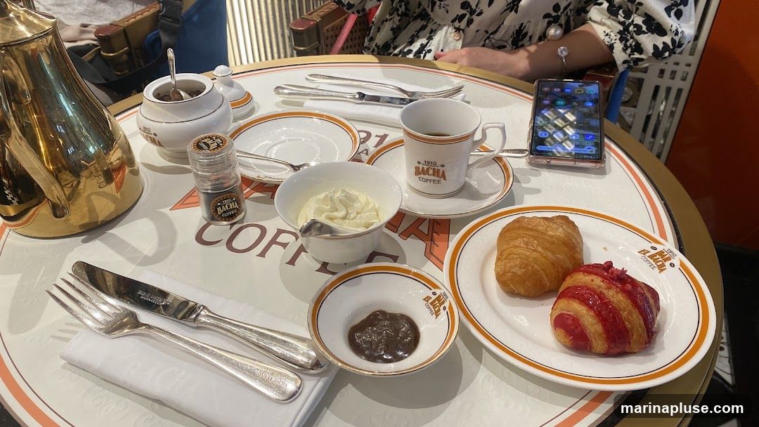 A table set for breakfast at Bacha Coffee, featuring a golden teapot, a cup of coffee, a bowl of cream, a small dish of jam, and two pastries on a plate. The table is adorned with the Bacha Coffee logo. A smartphone is also visible on the table.